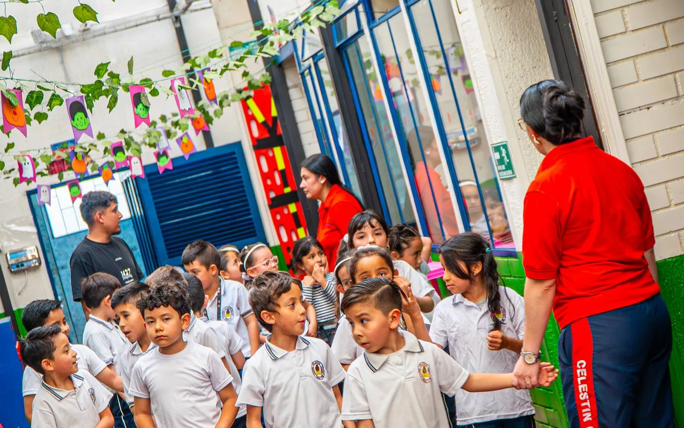 Niños y niñas con uniforme blanco recorren un pasillo decorado con guirnaldas de hojas y banderines; los acompañan varios adultos y una mujer con camiseta roja lleva de la mano a un niño.