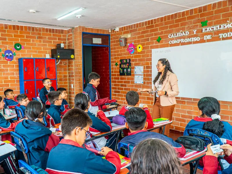 Salón con paredes de ladrillo y casilleros rojos; una docente sonríe junto al tablero blanco mientras estudiantes con uniforme azul y rojo la escuchan desde sus pupitres.