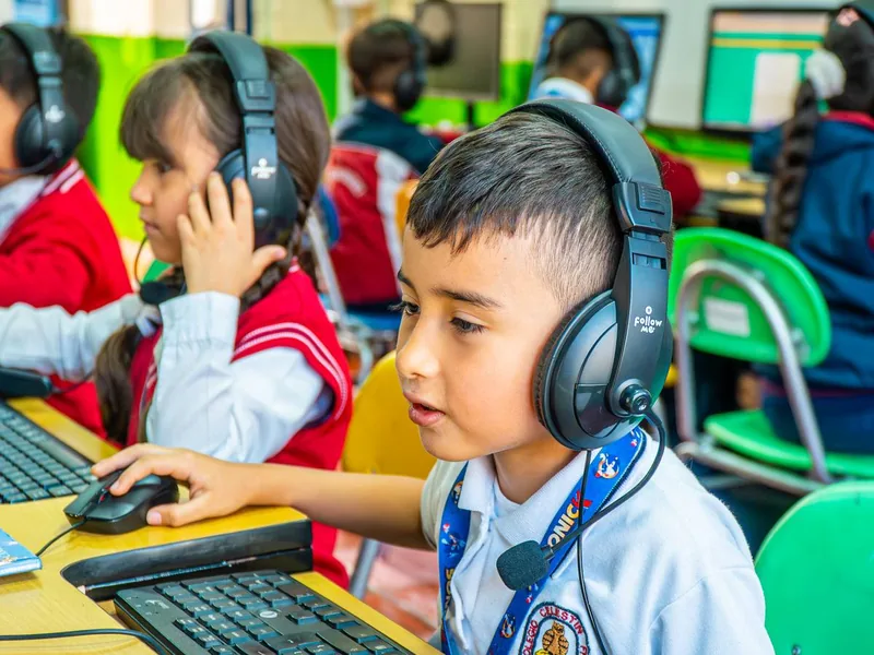 Un niño con audífonos y micrófono usa el mouse frente a un computador en una sala de cómputo; a su lado otros niños con audífonos trabajan en sus equipos.