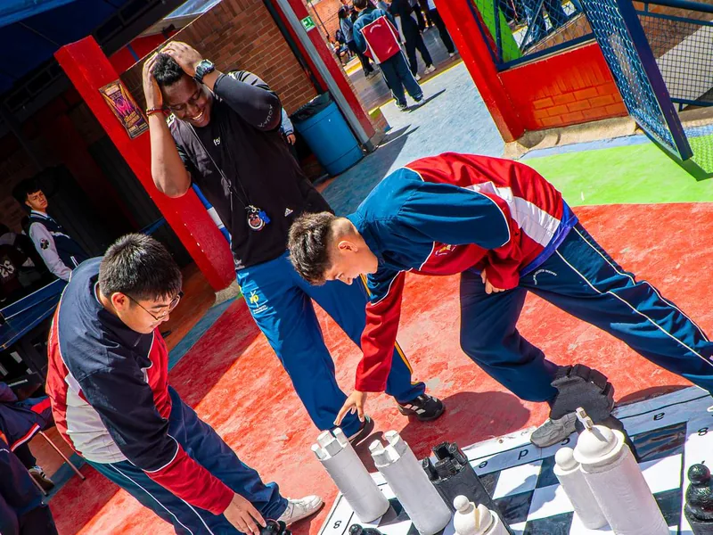 Dos estudiantes con uniforme deportivo se inclinan para mover fichas en un tablero de ajedrez gigante pintado en el piso, mientras un adulto se ríe con las manos en la cabeza.
