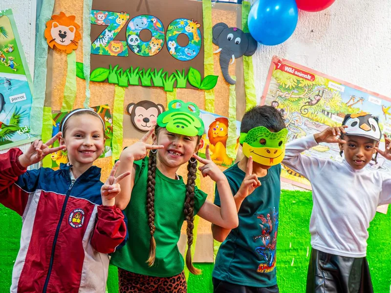 Cuatro niños sonríen y hacen la señal de paz frente a un mural con la palabra ZOO y carteles de animales con palabras en inglés; tres de ellos llevan antifaces de animales.