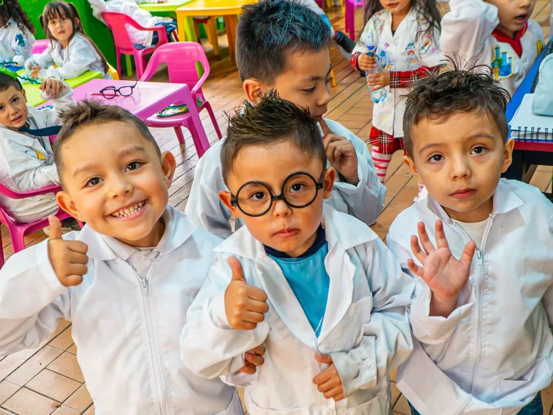 Tres niños pequeños con batas blancas de laboratorio posan en un salón de mesas de colores; uno sonríe con el pulgar arriba, otro lleva gafas redondas y otro saluda con la mano.