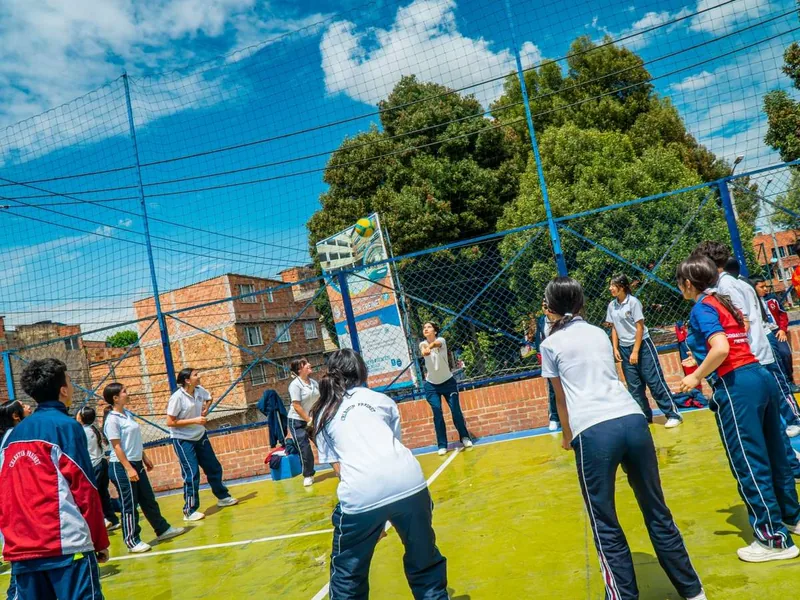 Estudiantes juegan voleibol en una cancha al aire libre rodeada de malla, con árboles grandes, cielo azul y edificios de ladrillo al fondo.