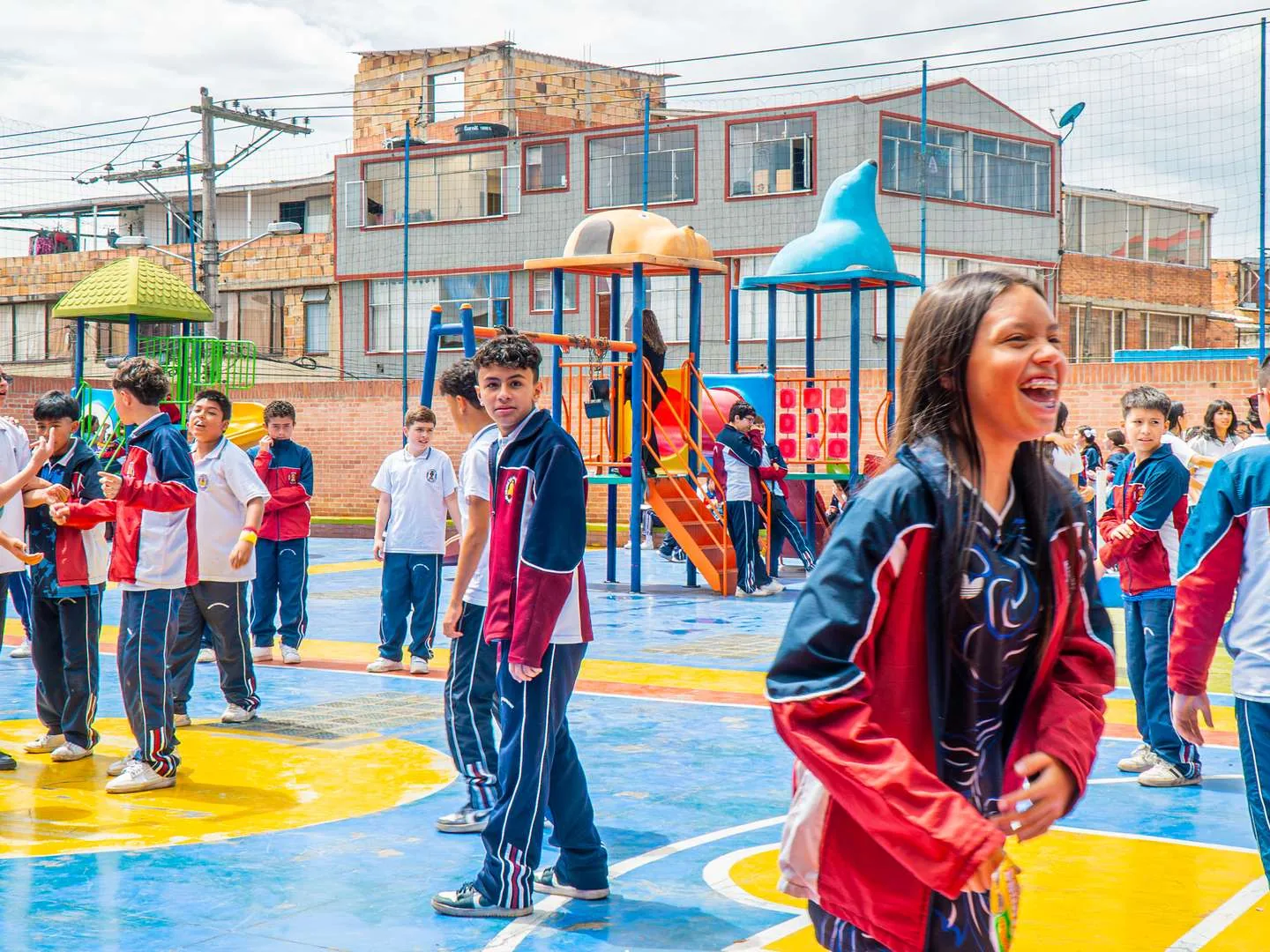 Estudiantes de bachillerato con uniforme azul y rojo en una cancha pintada de colores; en primer plano una estudiante ríe a carcajadas y al fondo se ven un parque infantil, mallas y edificios de ladrillo del barrio.