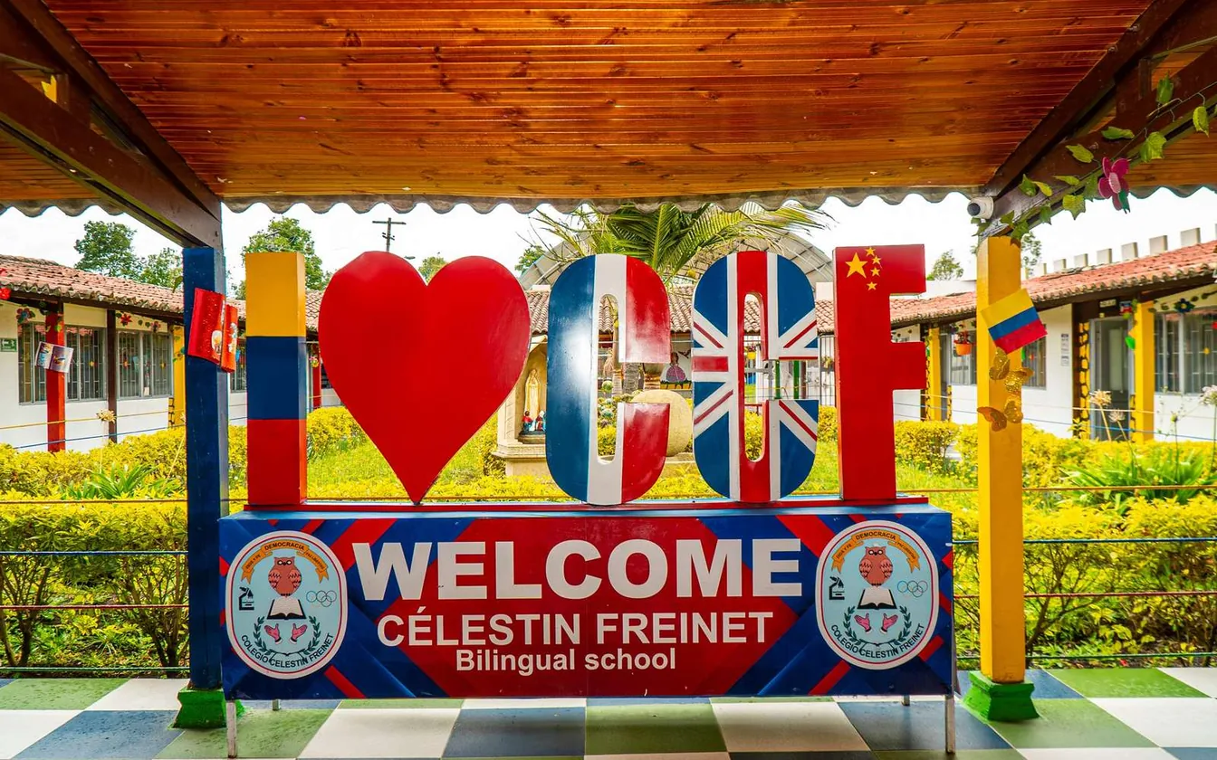 Letrero de bienvenida con las letras CCF, un corazón y banderas, que dice Welcome Célestin Freinet Bilingual school, bajo un techo de madera.
