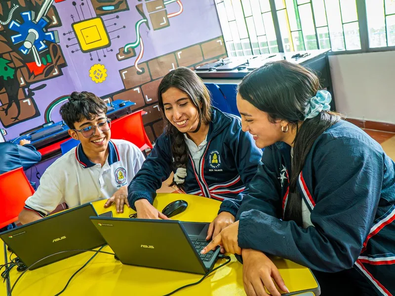 Tres estudiantes de bachillerato sonríen mientras trabajan en computadores portátiles en una sala con un mural de robots.