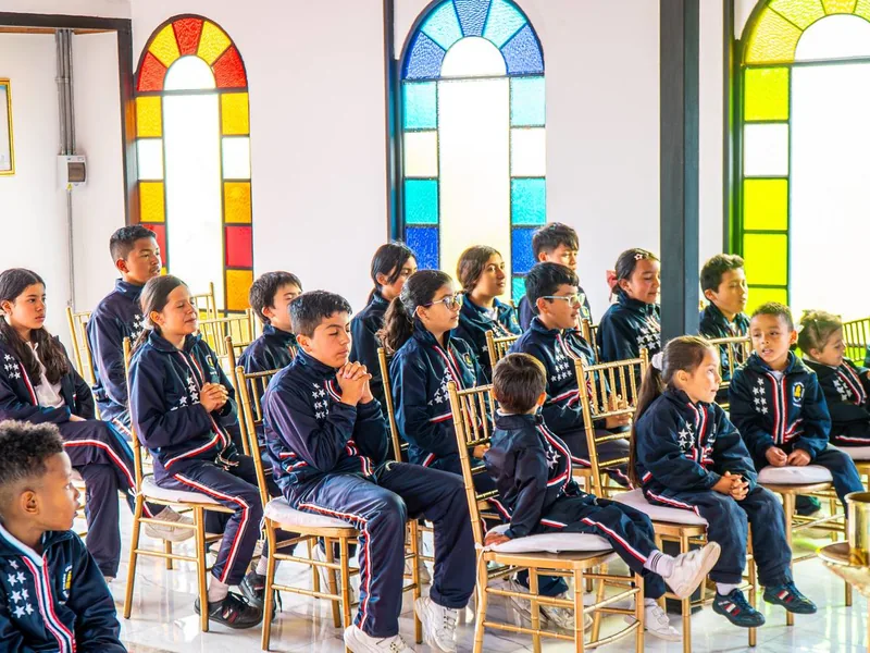 Estudiantes con uniforme azul sentados en sillas doradas dentro de una capilla con vitrales de colores.