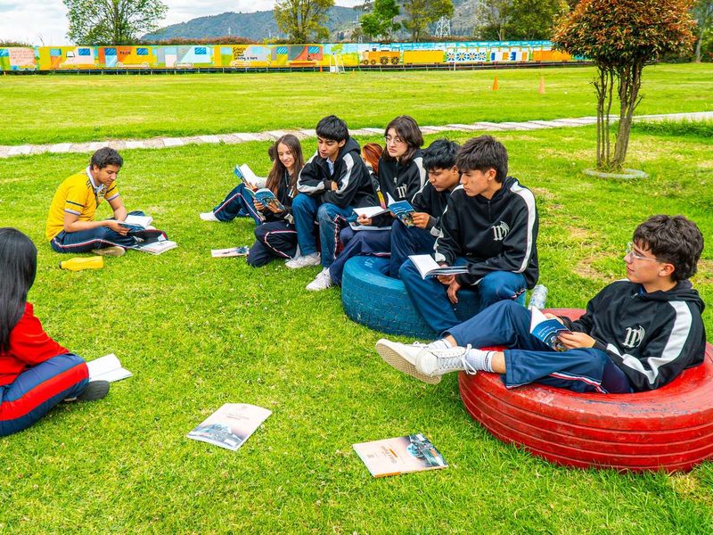 Estudiantes de bachillerato leen sentados en el césped y sobre llantas pintadas, con zonas verdes y montañas al fondo.