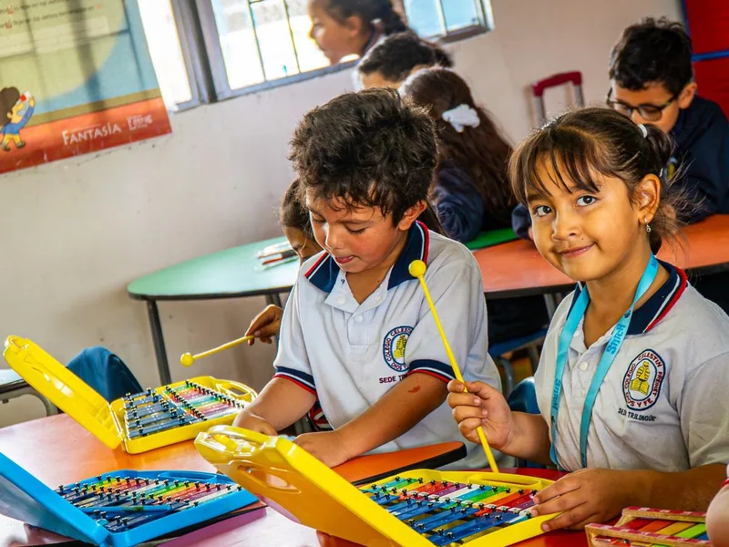 Una niña sonríe mientras toca un xilófono junto a un compañero en un salón de colores.