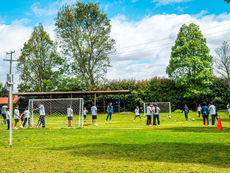 Estudiantes juegan fútbol en una amplia cancha de césped rodeada de árboles altos y cielo azul.