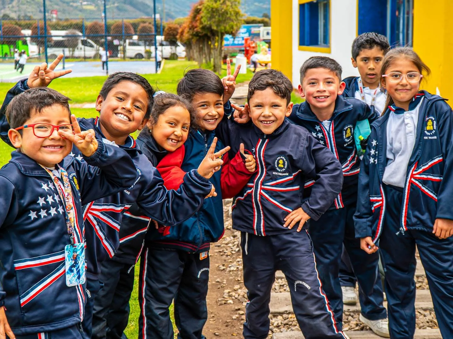 Niños de primaria con chaqueta azul del uniforme sonríen y hacen la señal de paz junto a la fachada amarilla y blanca del colegio, en una zona verde.
