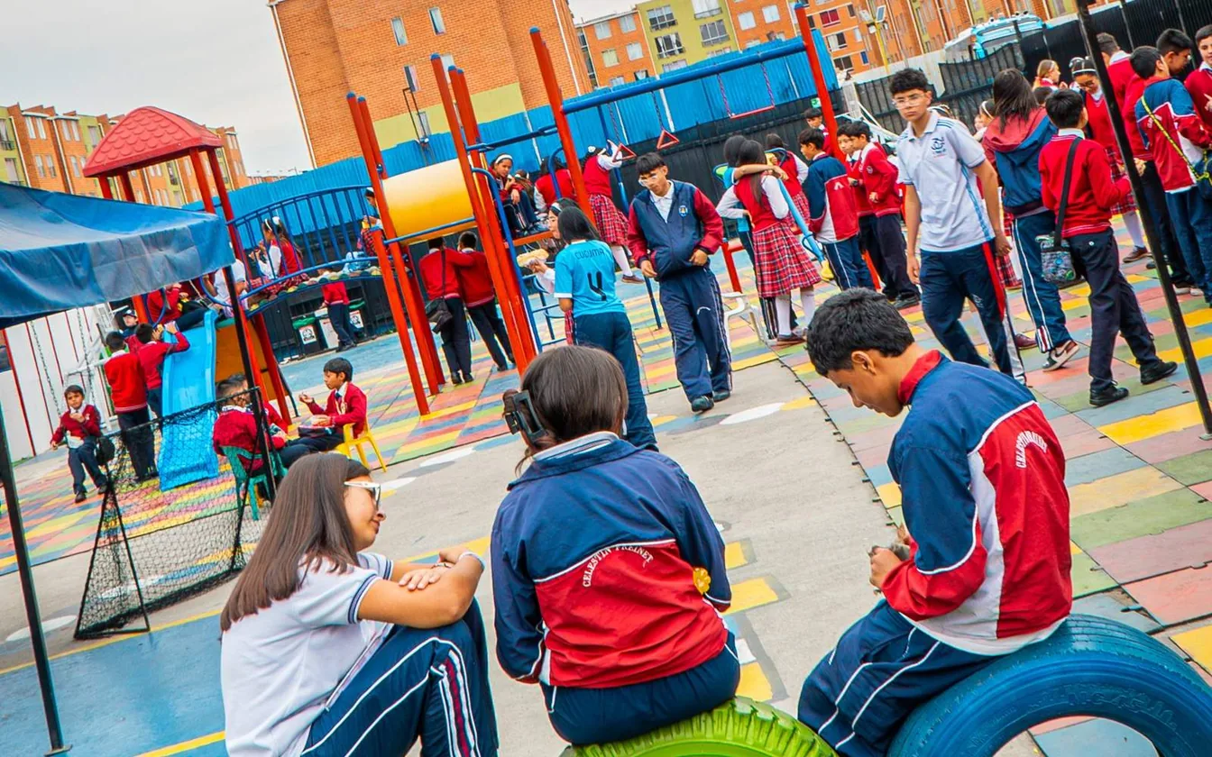 Estudiantes en un patio con juegos infantiles de colores, llantas pintadas y los edificios de ladrillo de Ciudad Verde al fondo.
