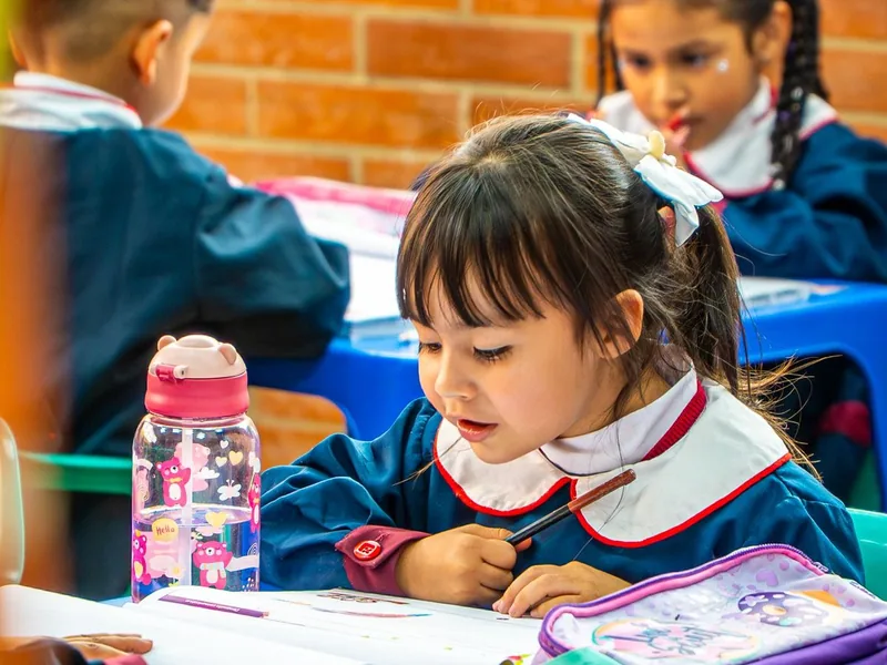 Una niña de preescolar dibuja concentrada en su pupitre junto a sus compañeros.