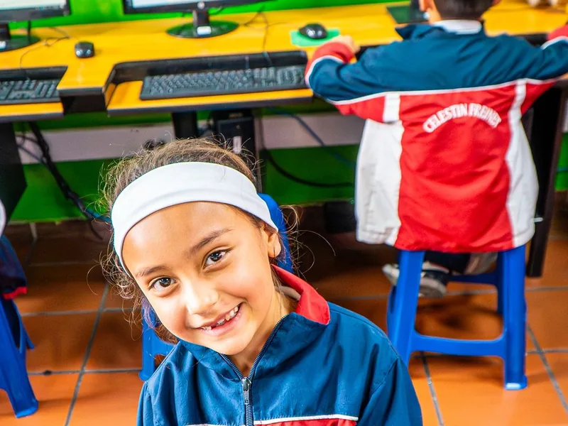 Una niña con diadema blanca sonríe en la sala de cómputo; detrás, un compañero trabaja frente a un computador.
