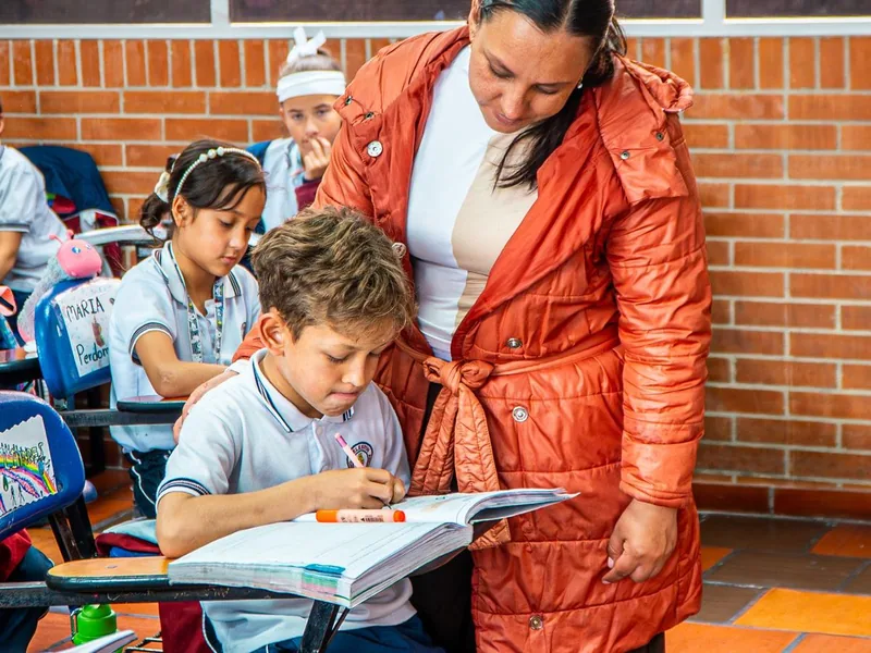 Una adulta acompaña a un estudiante que escribe en su cuaderno dentro del salón.