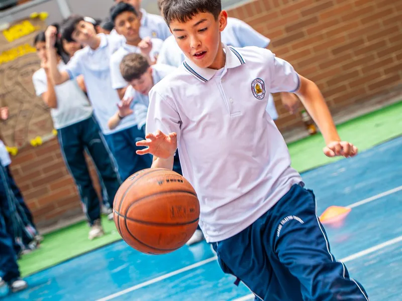 Un estudiante con uniforme de educación física corre con un balón de baloncesto en una cancha azul.