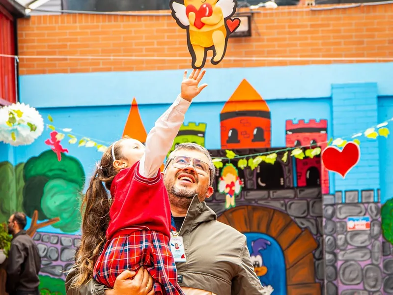 Un adulto del colegio levanta en brazos a una niña sonriente frente a un mural de castillo en la zona de preescolar.
