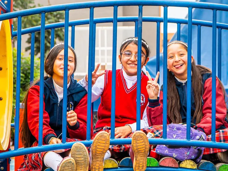Tres estudiantes sonríen sentadas en un juego del patio y hacen la señal de paz.