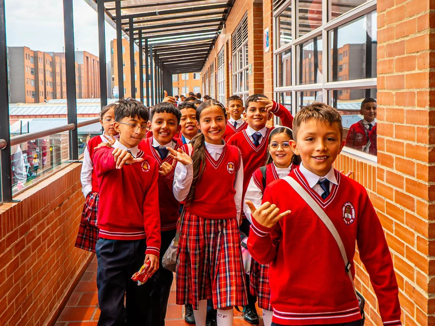 Estudiantes con uniforme rojo caminan sonrientes por un pasillo de ladrillo con ventanales; al fondo se ven los edificios de la ciudadela.