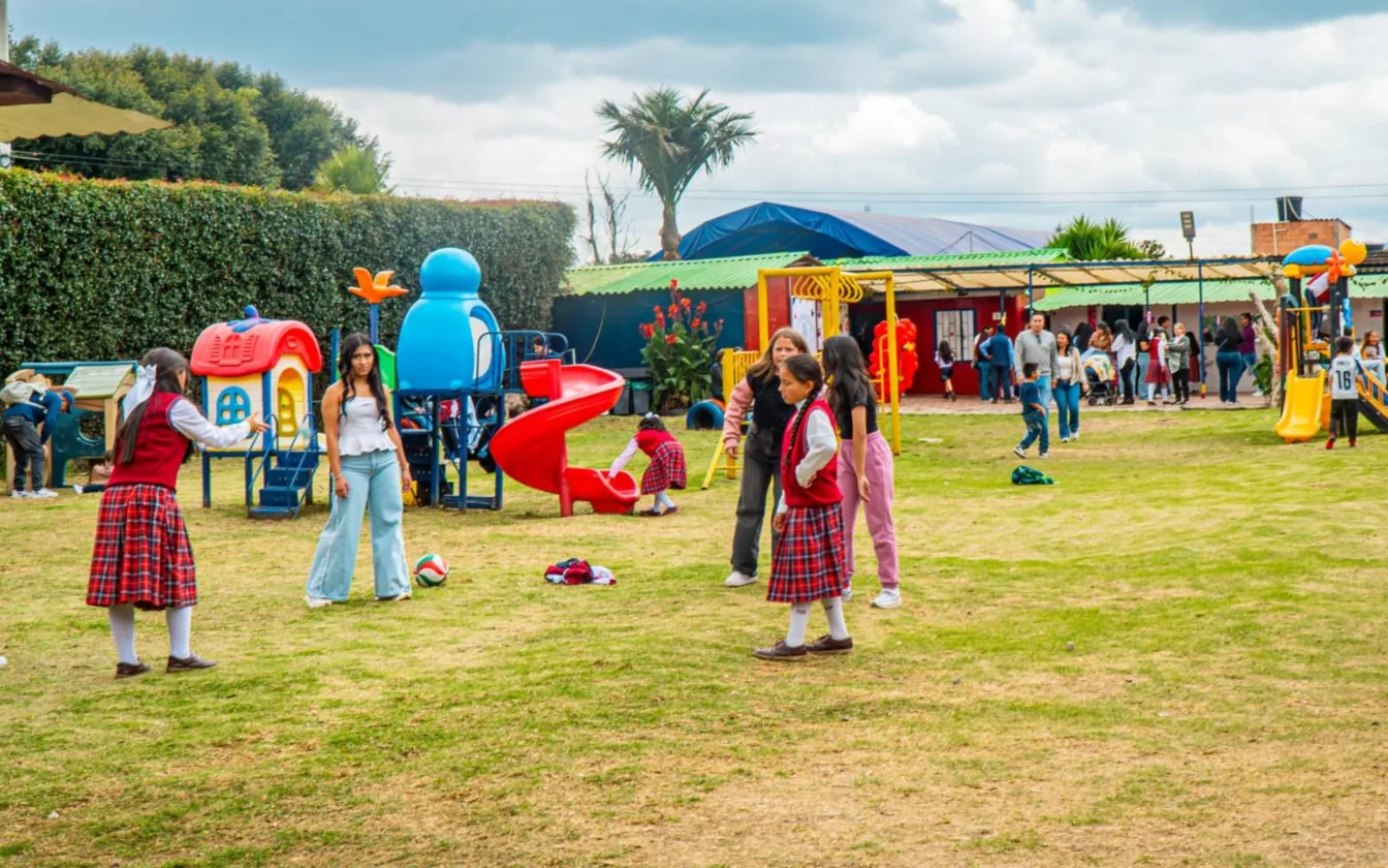 Estudiantes y adultos caminan por una amplia zona verde con juegos infantiles de colores y árboles al fondo.