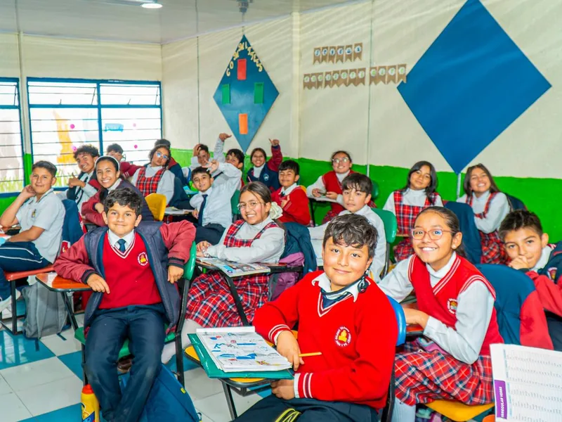 Estudiantes con uniforme de diario sonríen sentados en sus pupitres dentro de un salón decorado.