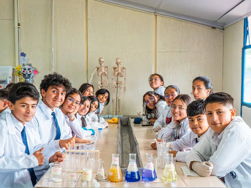Estudiantes con bata blanca sonríen alrededor de una mesa de laboratorio con frascos de líquidos de colores.
