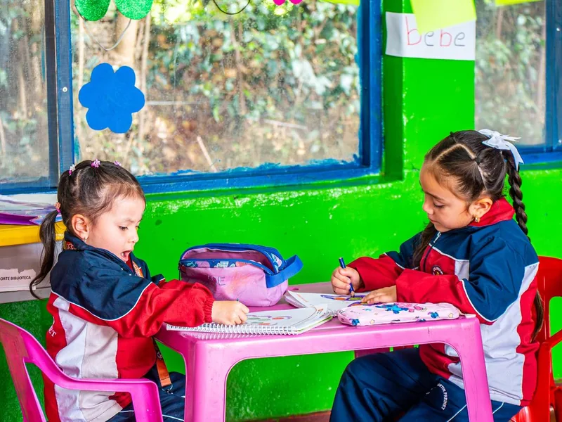 Dos niñas de preescolar dibujan en una mesa rosada dentro de un salón de colores.