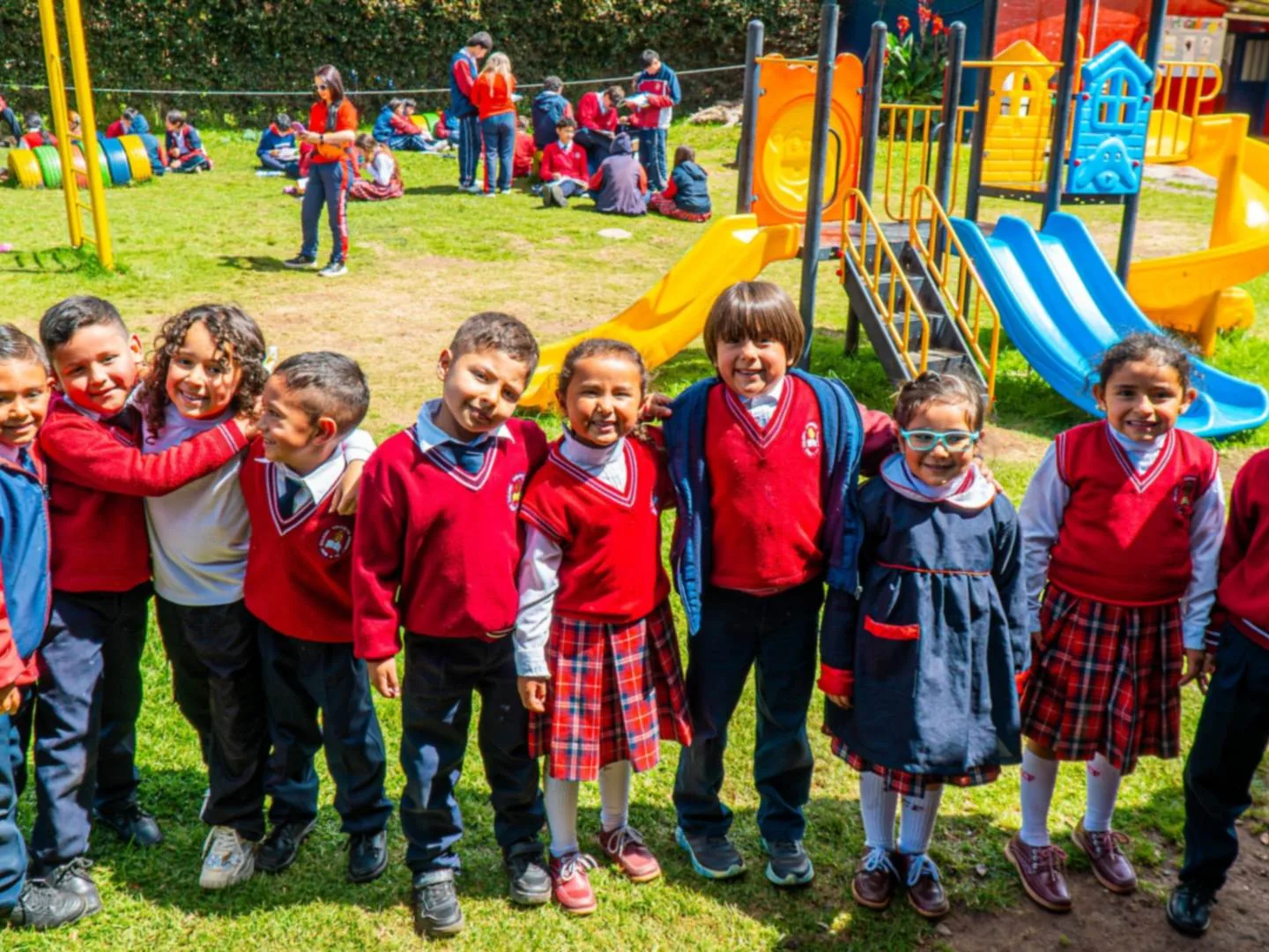 Niños y niñas de primaria con uniforme rojo y azul sonríen abrazados en una zona verde, con un parque infantil al fondo.